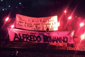 "Hurry to attack capital before a new ideology makes it sacred to you," reads a banner commemorating Alfredo's life. Hoisted by numerous masked figures holding flares in the night.